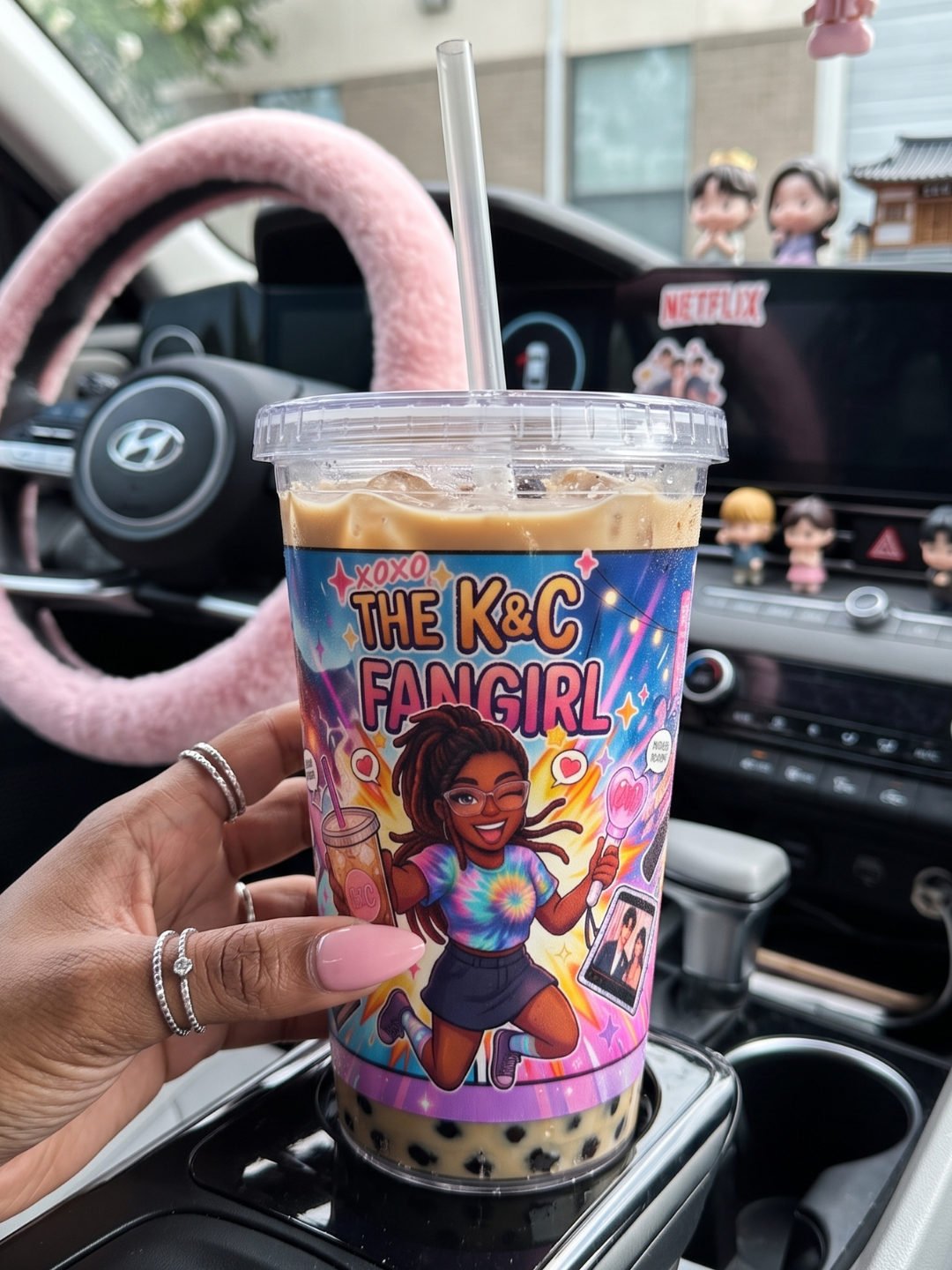 The K&C Fangirl Signature Comic Wrap 16oz Tumbler held in a car cup holder, filled with iced coffee and boba pearls, showcasing the colorful 2D webtoon pop-art character design.